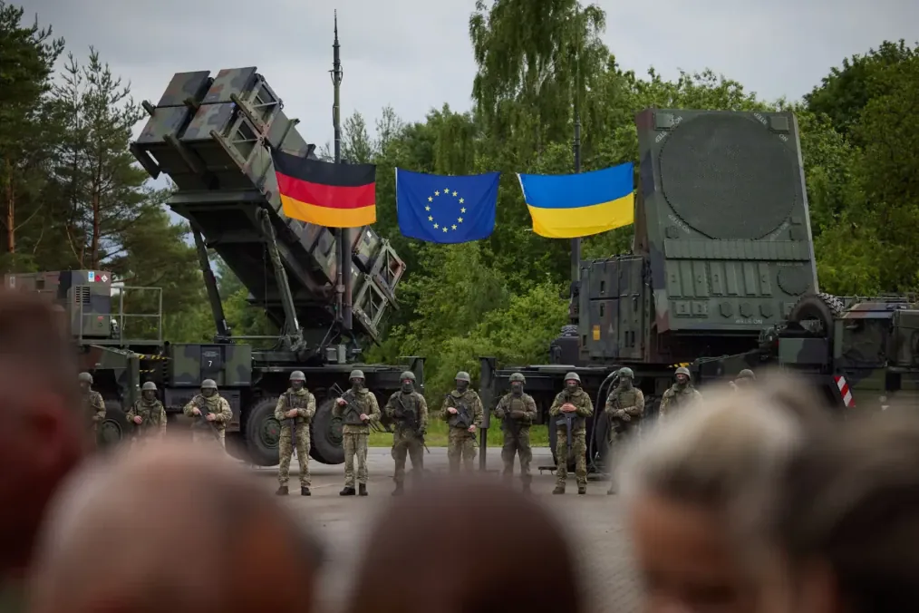 Ukrainian service members stand in front of a Patriot air defense system alongside German, EU, and Ukrainian flags. Ukrainian service members stand in front of a Patriot air defense system alongside German, EU, and Ukrainian flags.