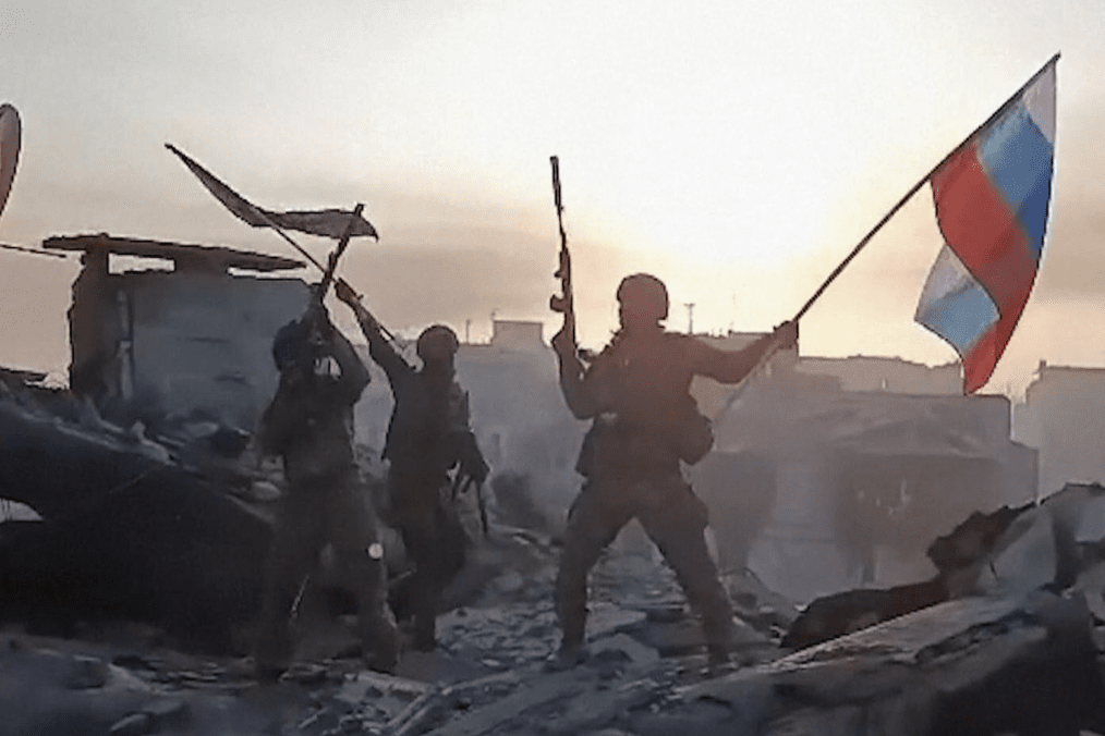Russian soldiers pose with a Russian flag amid destroyed buildings in footage published online. (Photo: Open source)
