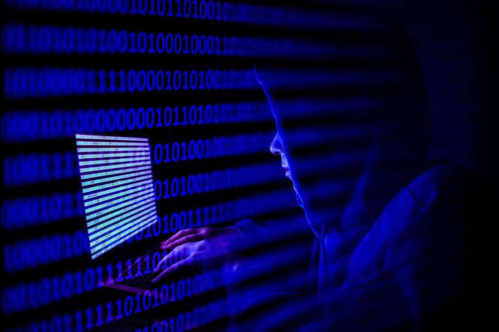 A person dressed as an internet hacker is seen with binary code displayed on a laptop screen. (Source: Getty Images)