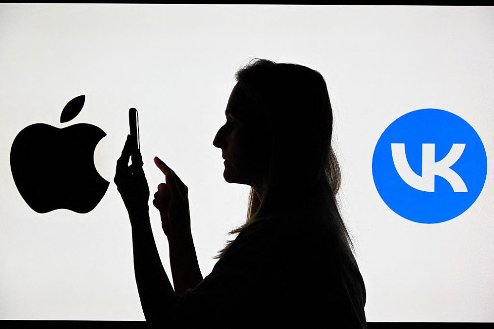 This illustration photograph shows a woman using a smartphone in front of the logos of Russian social media platform VK and Apple Inc displayed on a TV screen. (Source: Getty Images)