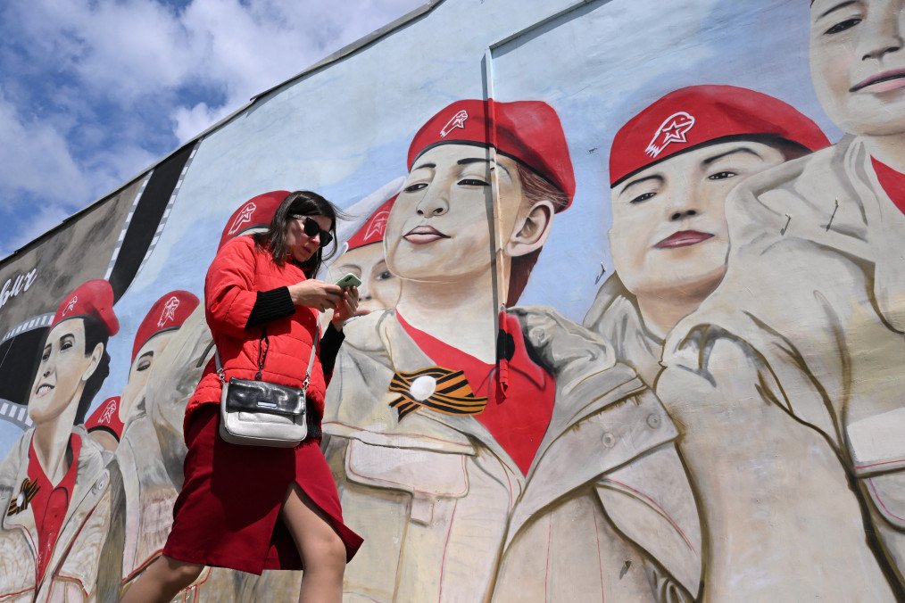 A woman uses her smartphone as she walks past a mural depicting members of Russia's Yunarmiya (Young Army) patriotic youth movement in Moscow on May 14, 2024.