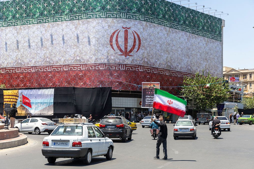 A man is seen waving an Iranian flag in front of a billboard displaying the Iranian flag on June 15, 2026 in Tehran, Iran. (Source: Getty Images)