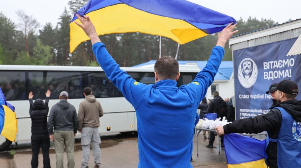 Tres soldados ucranianos delgados abrazados y cubiertos con banderas azules y amarillas en un campo abierto durante el proceso de intercambio de prisioneros.