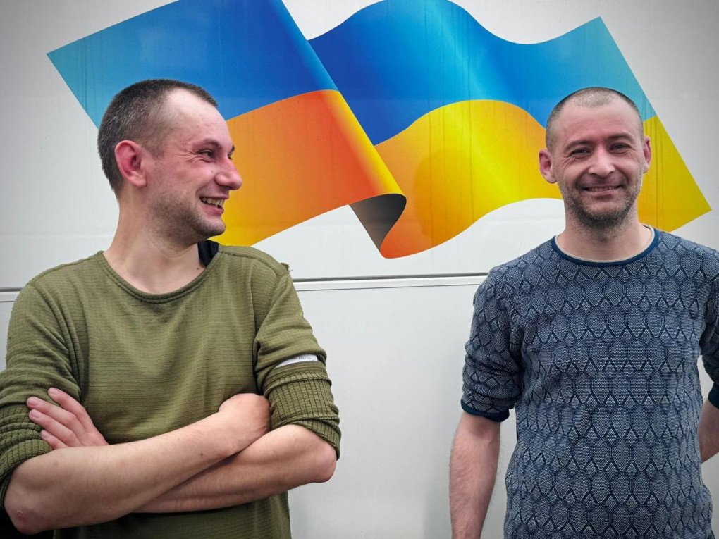 Two released Ukrainians smile after returning from captivity, with the national flag in the background. (Source: Volodymyr Zelenskyy / Telegram)