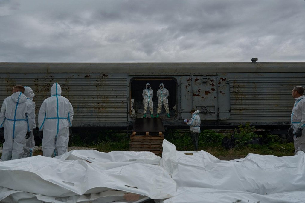 Ukrainian forensic experts in full white protective gear. Several dozen white body bags are arranged in neat rows on the ground in the foreground. Two experts stand in the open doorway of the refrigerated car, which is filled with more remains returned from Russia. Other experts on the ground take notes and inspect the bags as part of a large-scale identification effort.