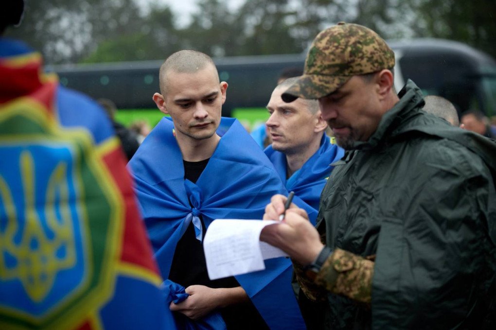 Tres soldados ucranianos delgados abrazados y cubiertos con banderas azules y amarillas en un campo abierto durante el proceso de intercambio de prisioneros.