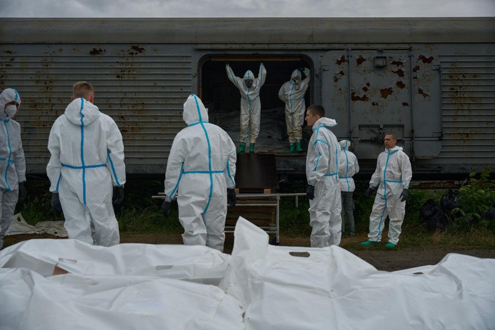 Ukrainian forensic experts in full white protective gear. Several dozen white body bags are arranged in neat rows on the ground in the foreground. Two experts stand in the open doorway of the refrigerated car, which is filled with more remains returned from Russia. Other experts on the ground take notes and inspect the bags as part of a large-scale identification effort.