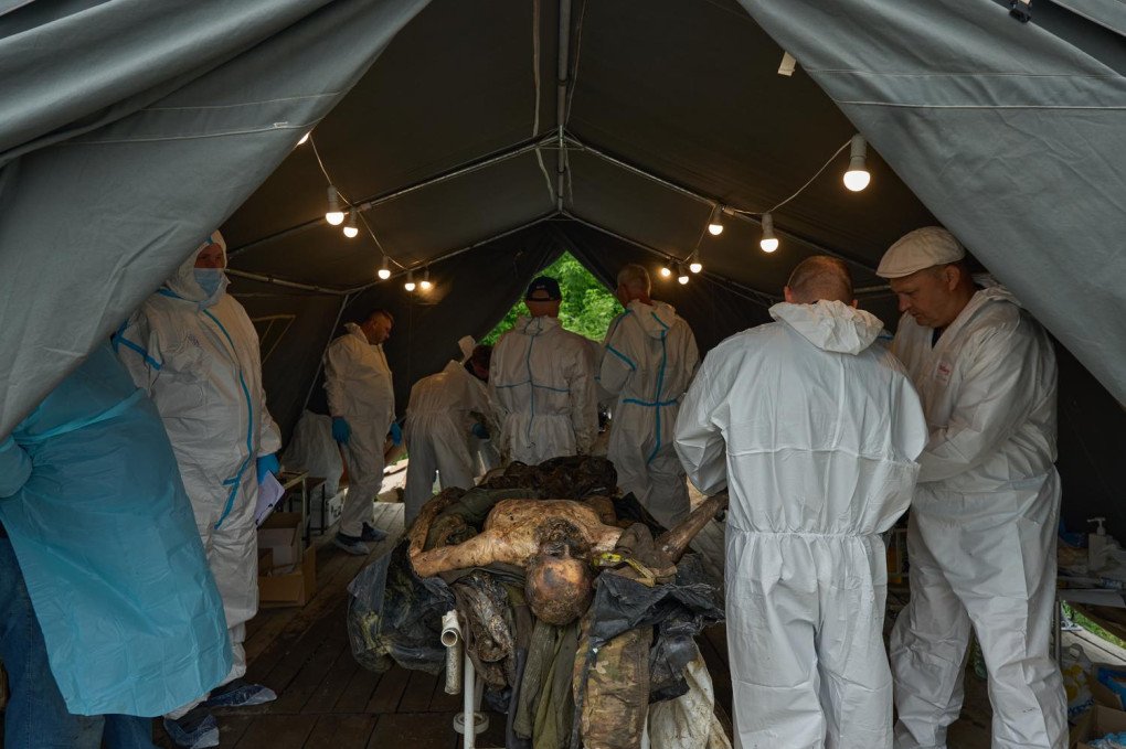 Inside a field tent under string lighting, several Ukrainian forensic experts wearing white hazmat suits and protective gear gather around an examination table. On the table lie the severely decomposed, mummified remains of a fallen soldier, with visible tatters of camouflage military uniform. The forensic specialists work methodically in groups, inspecting the body and documenting evidence to determine the cause of death and establish a DNA profile.