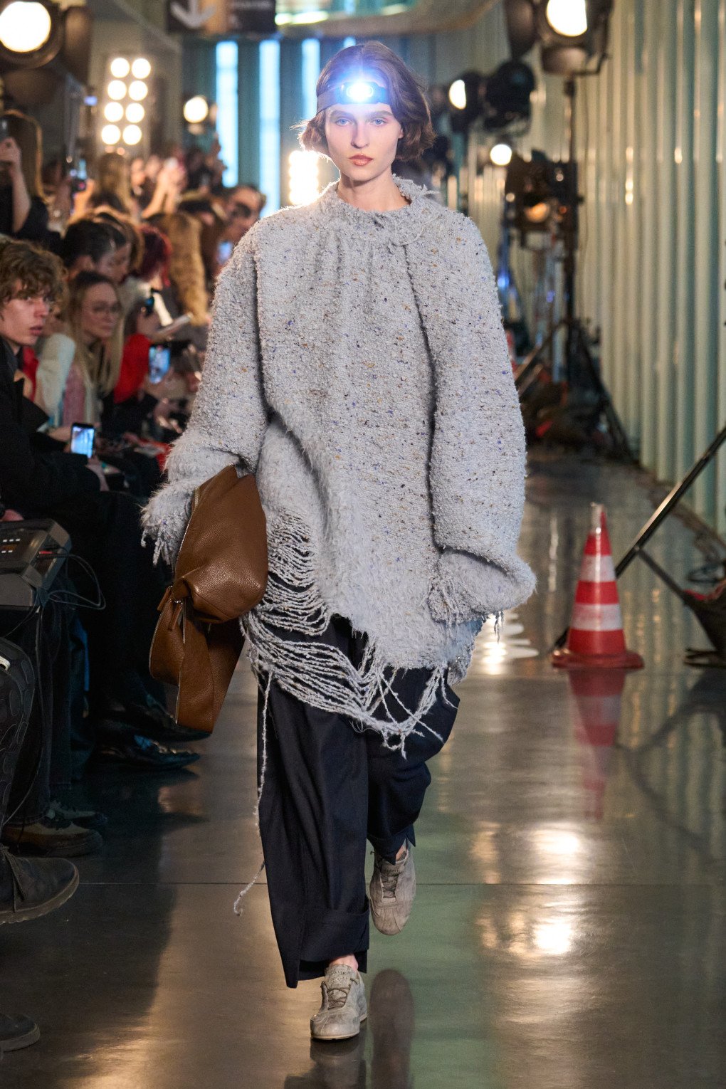 The model showcases a chunky grey knit sweater with frayed edges paired with dark loose pants and sneakers, a statement accessory being the headlamp lighting the way. (Source: LITKOVSKA press office)
