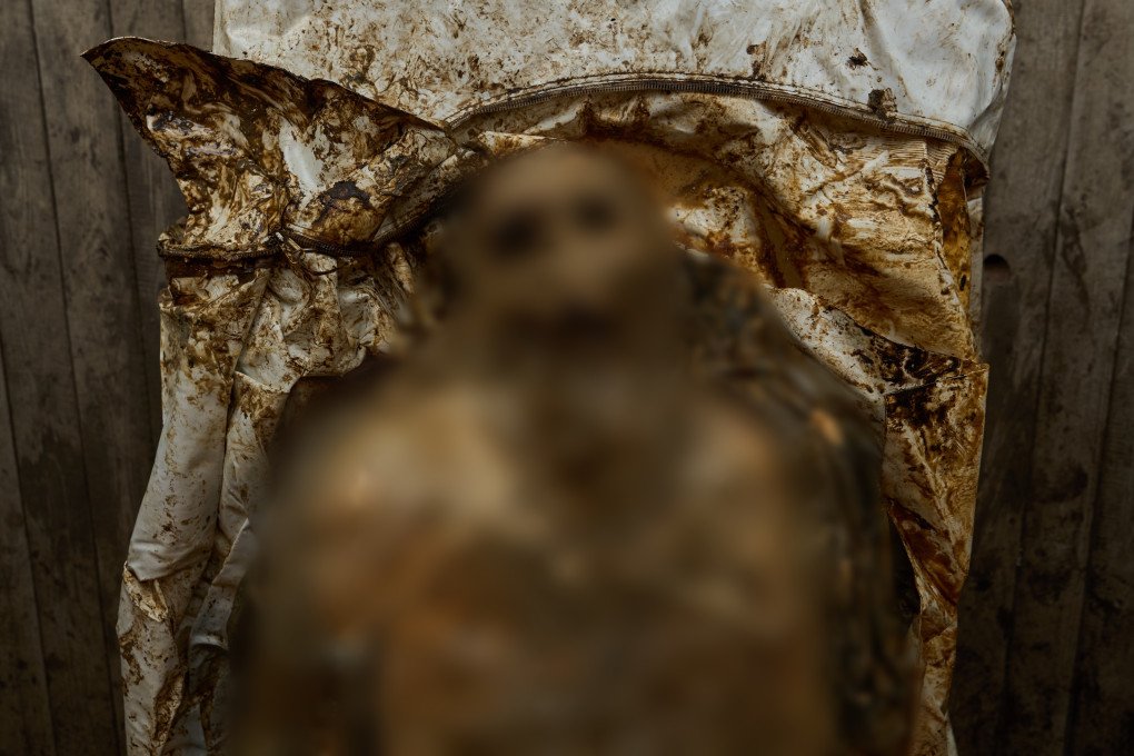 A close-up photograph shows the severely decomposed, mummified remains of a human body inside a soiled white body bag against a wooden background. The skin is deeply discolored, leathery, and decayed, exposing underlying facial bone structures, empty eye sockets, and teeth. Traces of a dark uniform are visible on the shoulders, illustrating the stark and brutal reality of frontline casualties awaiting forensic identification.