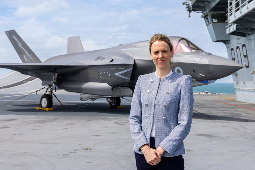 UK Minister for the Armed Forces Louise Sandher-Jones aboard a Royal Navy aircraft carrier, with an F-35B Lightning fighter jet in the background. (PHOTO: GOV.UK)