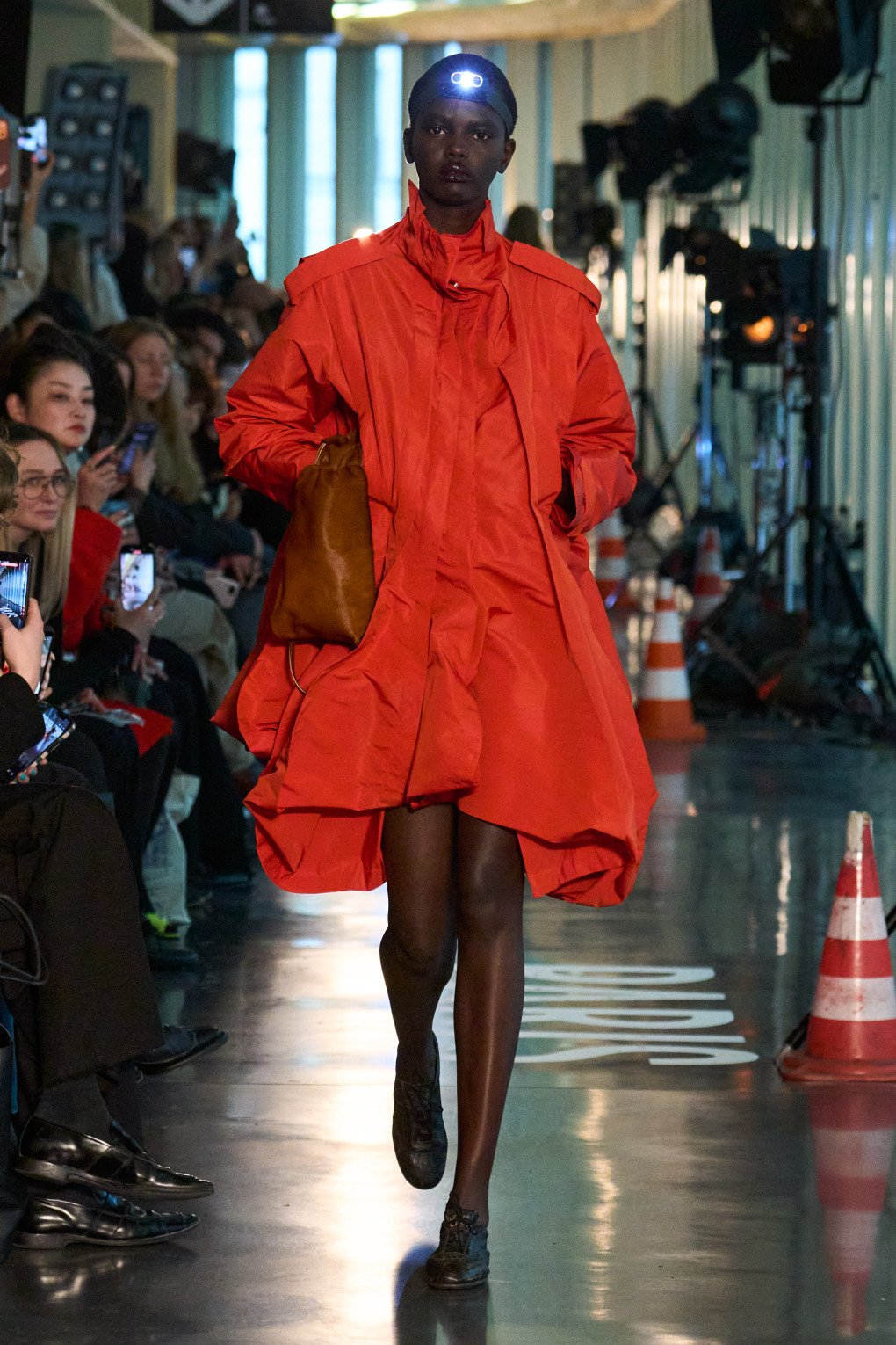 A model in a bold red oversized jacket with a dramatic silhouette walks confidently down the runway, carrying a brown leather bag, as the spotlight shines from behind. (Source: LITKOVSKA press office)