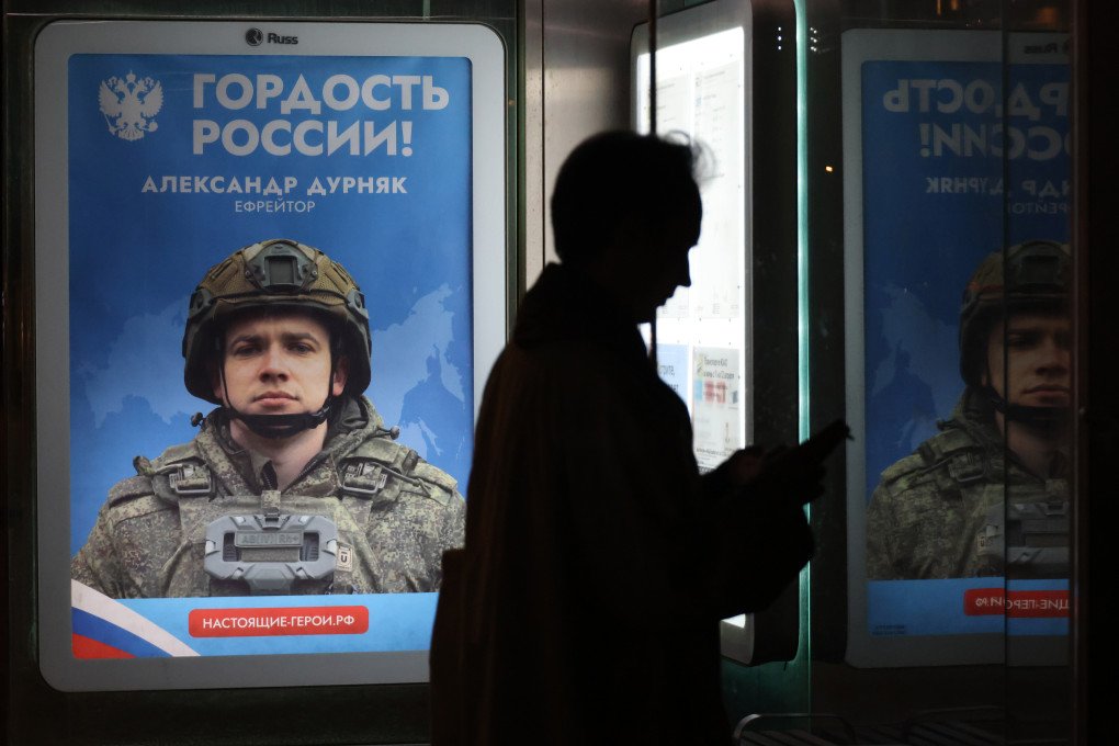 A woman holding a smartphone waits at a bus stop next to a propaganda banner depicting a Russian soldier invading Ukraine, on April 17, 2026, in Moscow, Russia. The banner reads, "Pride of Russia. Alexander Durnyak, Corporal. Real heroes". (Photo by Contributor via Getty Images)