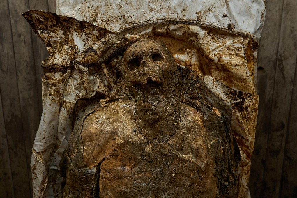 A close-up photograph shows the severely decomposed, mummified remains of a human body inside a soiled white body bag against a wooden background. The skin is deeply discolored, leathery, and decayed, exposing underlying facial bone structures, empty eye sockets, and teeth. Traces of a dark uniform are visible on the shoulders, illustrating the stark and brutal reality of frontline casualties awaiting forensic identification.