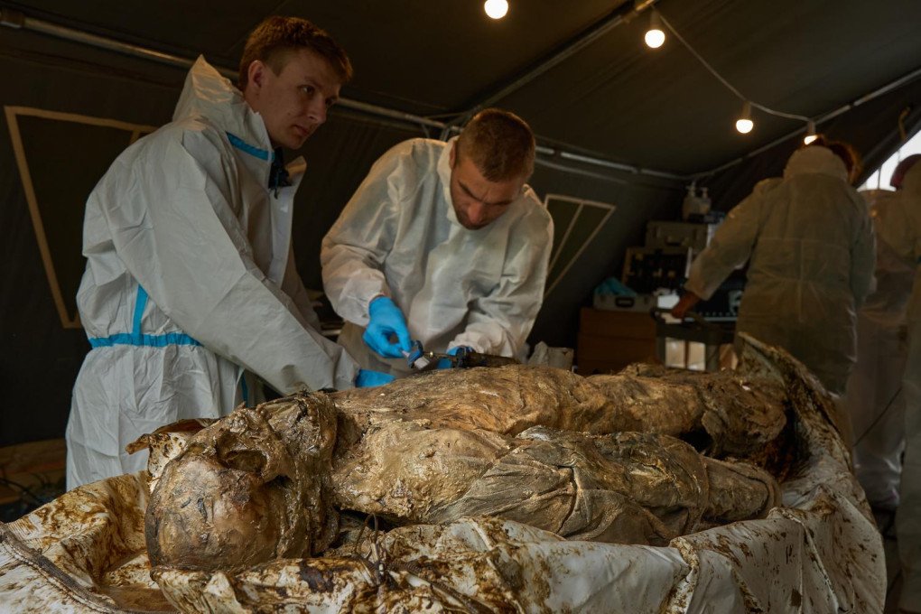 A close-up photograph shows two young male Ukrainian forensic specialists in white protective hazmat suits working intently over a severely mummified, decomposed human body. The expert on the right, wearing blue nitrile gloves, uses a specialized metal tool to carefully examine or stabilize the hand and fingers of the deceased soldier. The body is positioned on an examination table inside a temporary field tent lit by hanging incandescent bulbs.