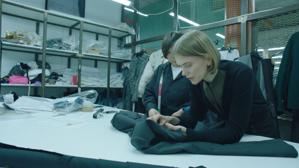 A designer carefully inspects the fabric, ensuring precision in their creation, amidst a busy workroom filled with materials and tools. (Source: press office)