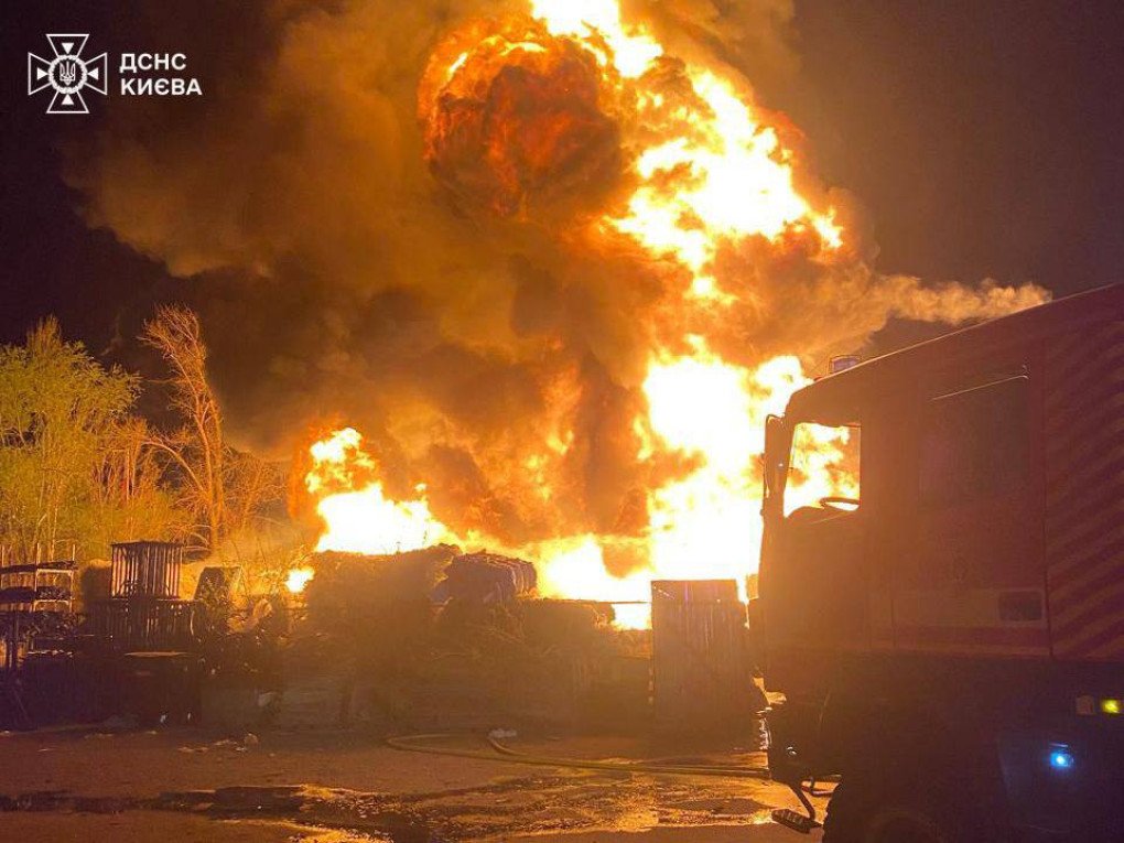 Bola de fuego masiva y espesa columna de humo elevándose sobre Kyiv durante la noche tras un impacto ruso, con camión de bomberos desplegado junto al incendio.