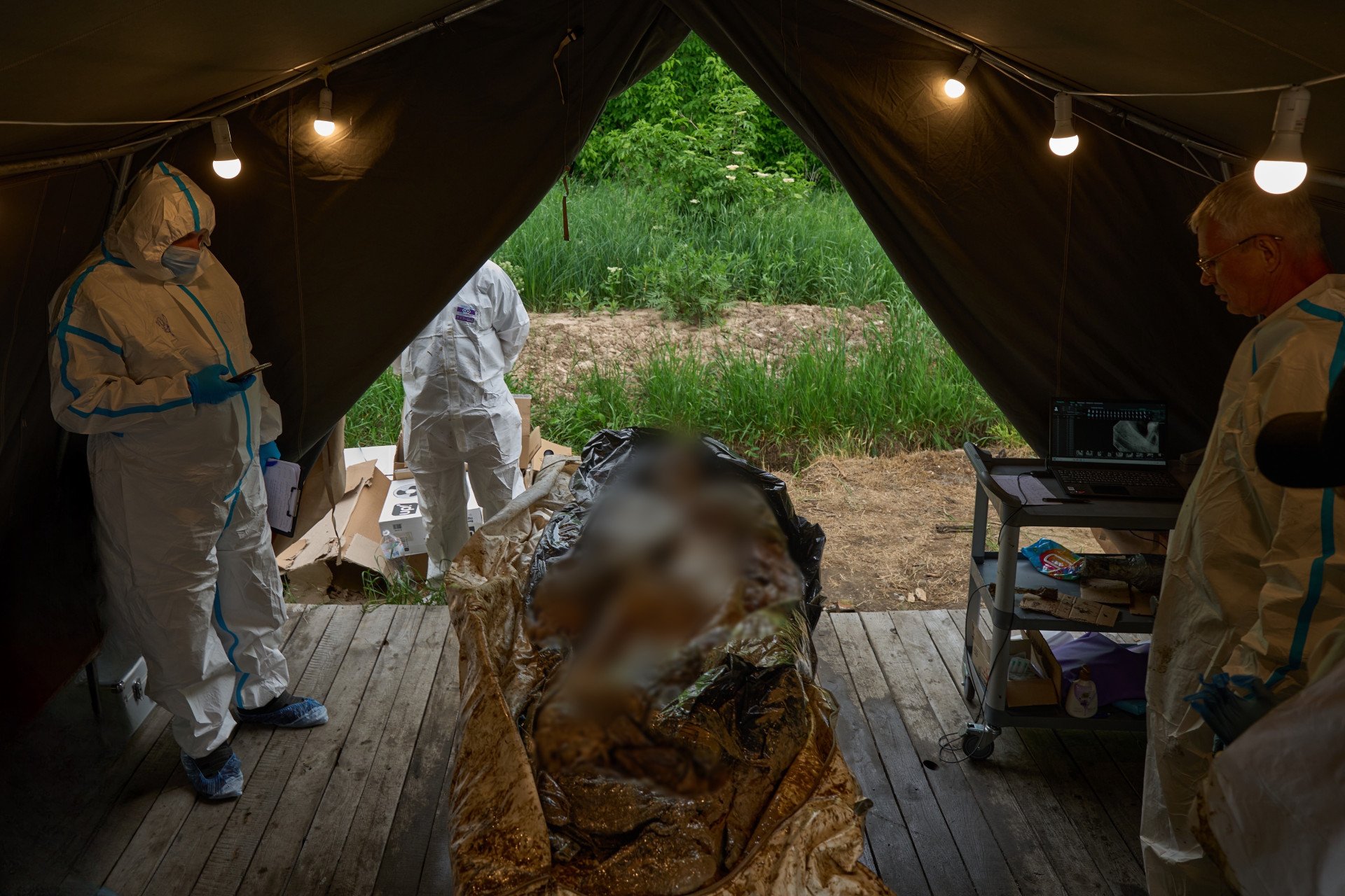 Inside a temporary field tent lit by hanging lightbulbs, Ukrainian forensic experts in white hazmat suits prepare to work on heavily decomposed human remains laid on a wooden floor. One expert stands on the left holding a smartphone and a clipboard, while another stands on the right near a mobile cart equipped with a laptop displaying a forensic image or scan. The open back of the tent looks out onto a green, grassy outdoor area.