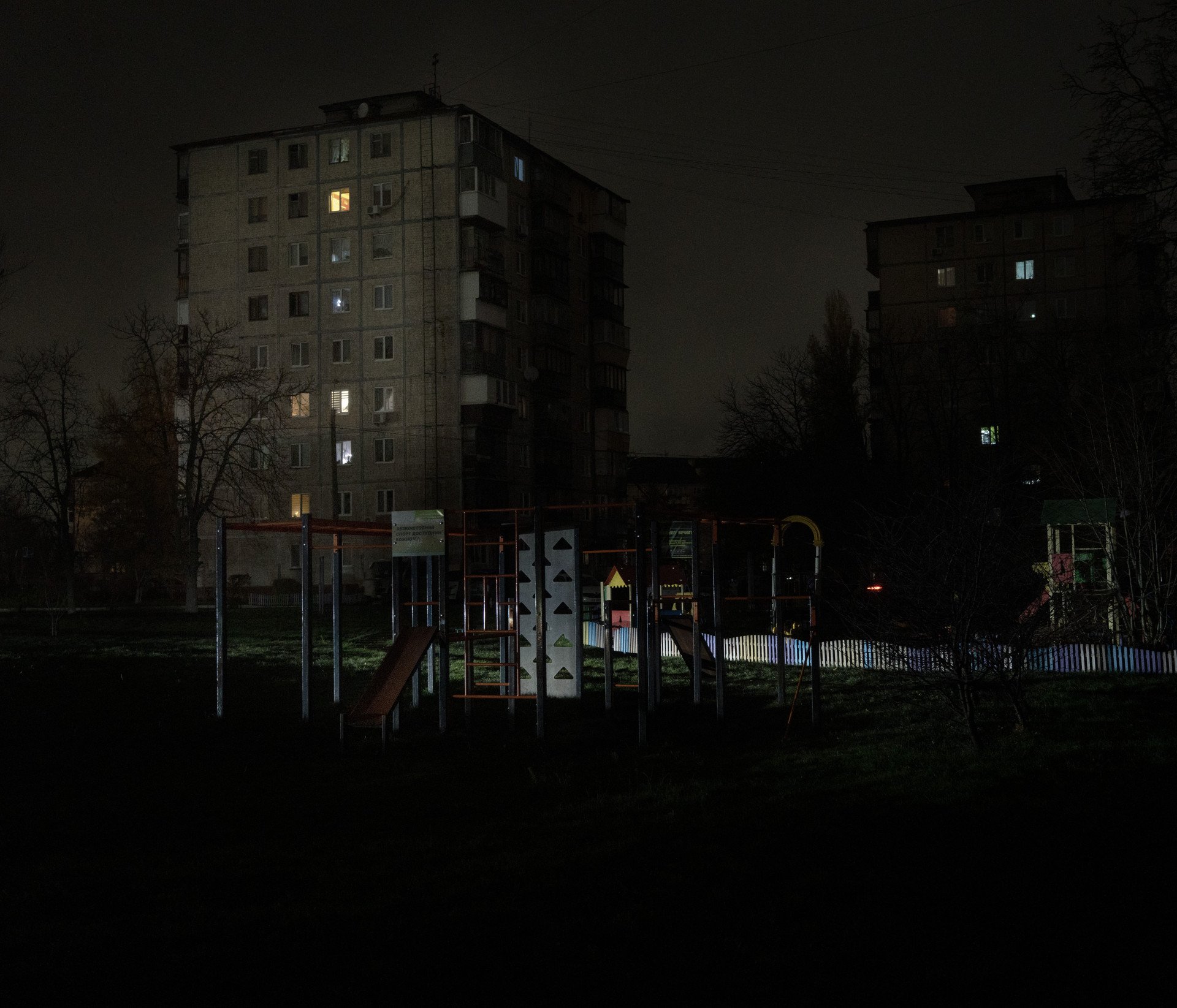 Un parque infantil resplandece bajo los faros de un coche que pasa, con edificios residenciales, en su mayoría oscuros, al fondo. 10 de noviembre de 2025. Foto de Joshua Olley/UNITED24 Media.