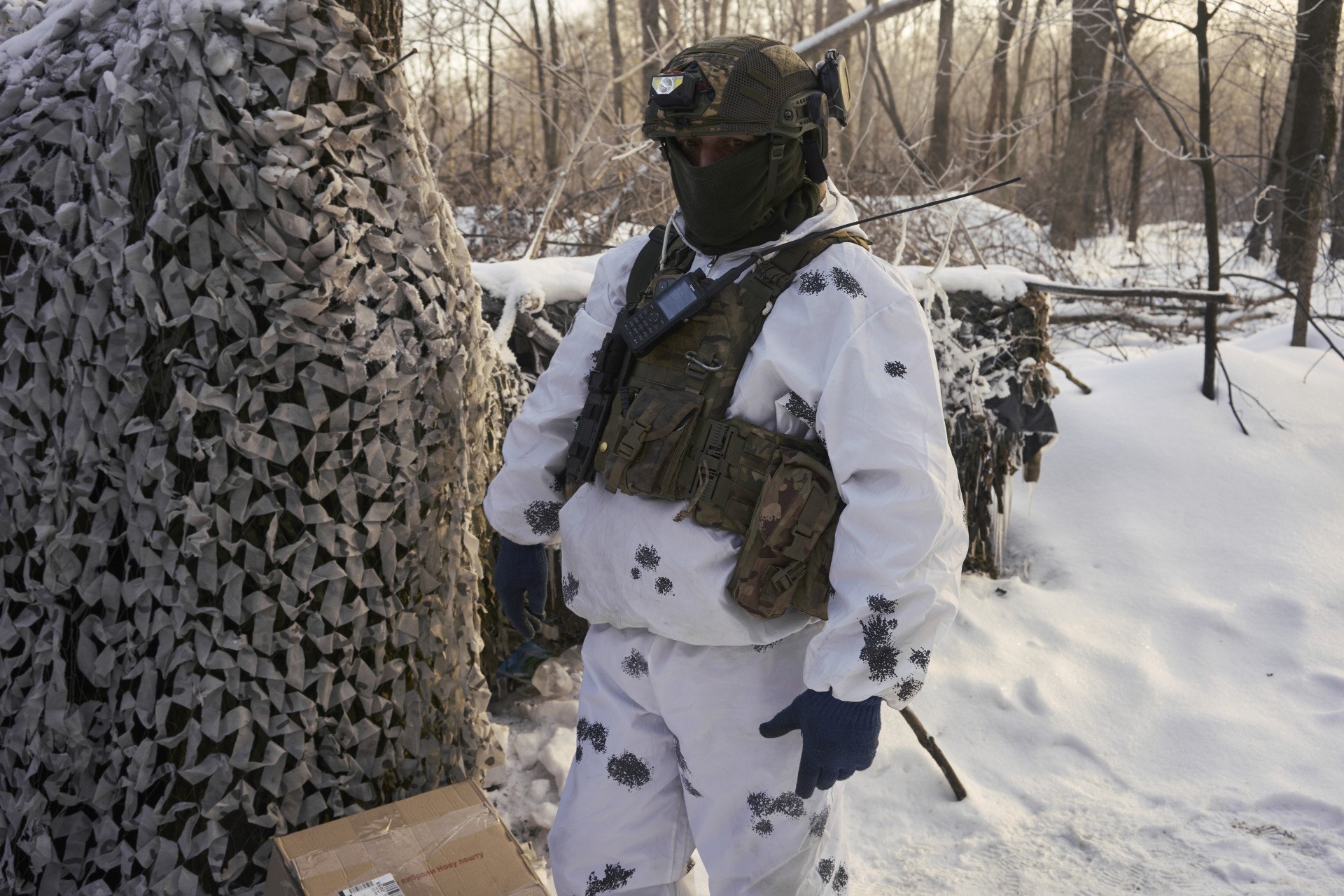 A Ukrainian servicemember wearing winter over-white camouflage stands beside a concealed position draped in netting, reflecting how frontline concealment increasingly depends on layered cover and silhouette disruption in snow-covered terrain, February 2026. (Photo: Joshua Olley/UNITED24) snow camo in Ukraine