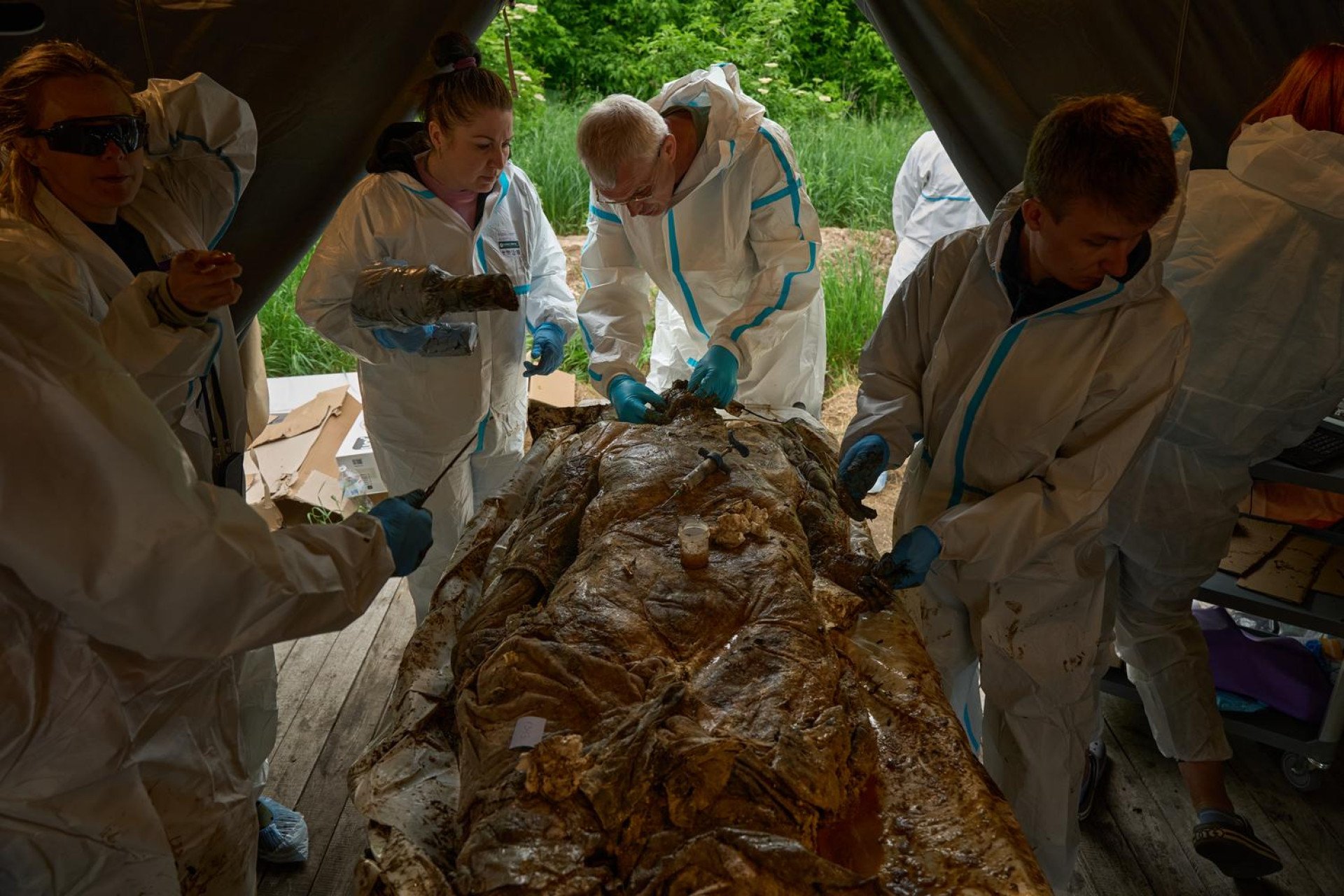 Inside a military field tent, a team of five Ukrainian forensic experts wearing white protective hazmat suits and blue nitrile gloves works around a heavily mummified, decomposed human body laid out on an examination table. In the center, an older male pathologist with glasses carefully inspects the oral and dental structures of the skull, while a female colleague assists from the left. Syringes and small sample containers rest on the chest of the deceased soldier during the post-mortem procedure.