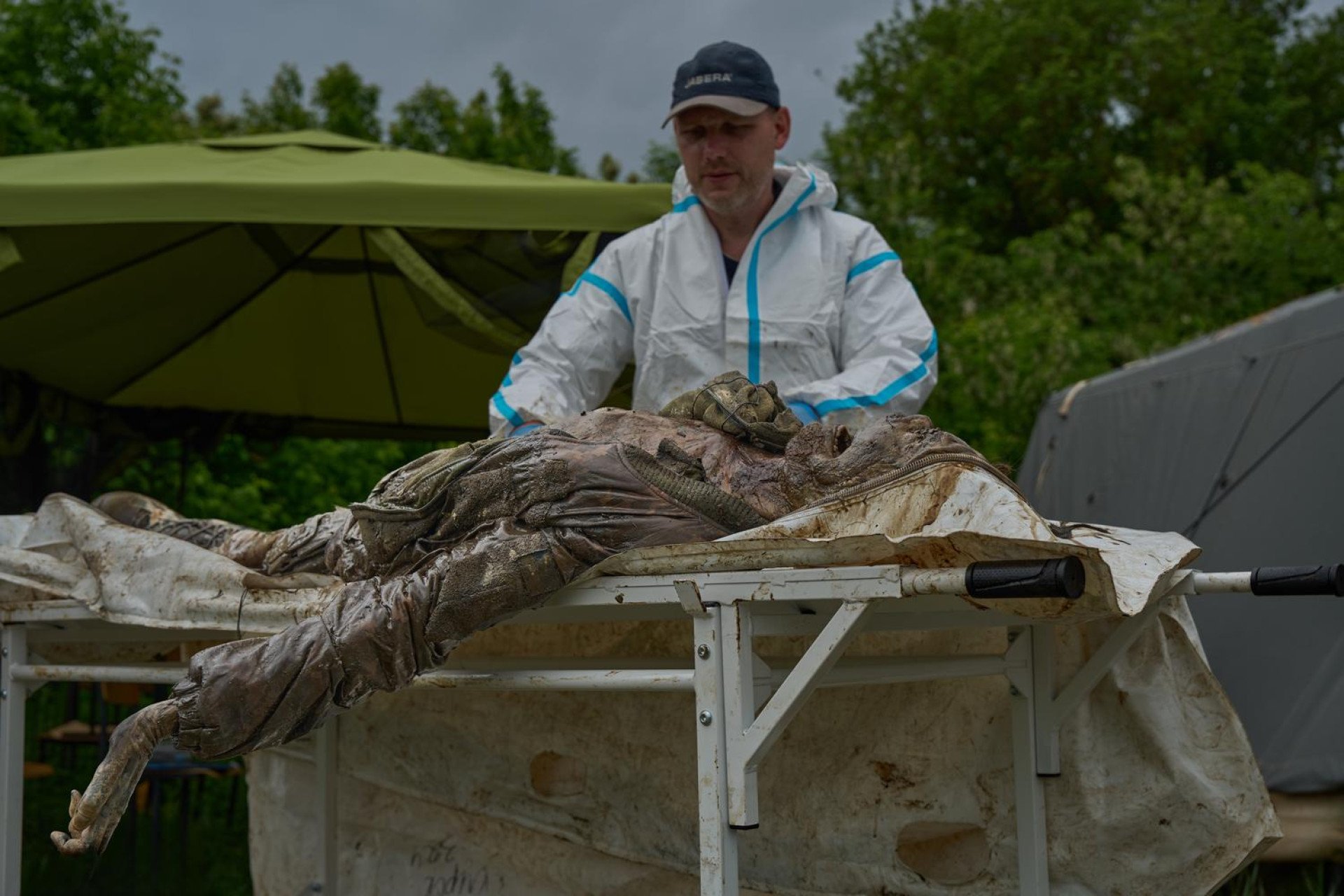 An outdoor photograph shows a Ukrainian forensic expert wearing a baseball cap and a white hazmat suit examining a body on a medical gurney. The heavily decomposed, dirt-covered remains are of a soldier still wearing remnants of tactical gear and combat boots. The expert works carefully near an open tent structure in the background, focusing on documenting trauma and identifying features on the remains.