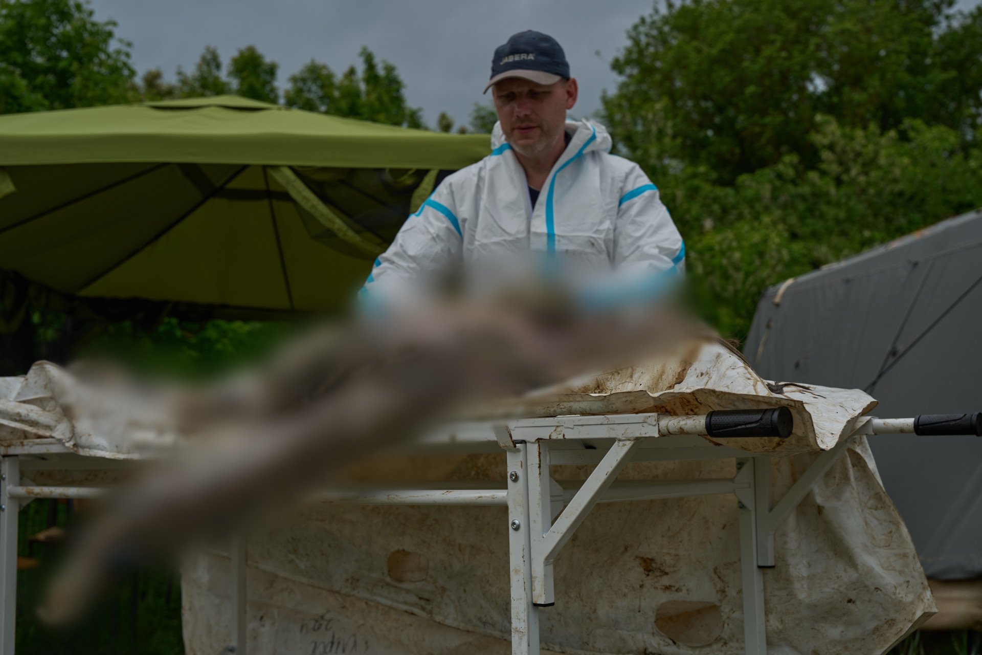 An outdoor photograph shows a Ukrainian forensic expert wearing a baseball cap and a white hazmat suit examining a body on a medical gurney. The heavily decomposed, dirt-covered remains are of a soldier still wearing remnants of tactical gear and combat boots. The expert works carefully near an open tent structure in the background, focusing on documenting trauma and identifying features on the remains.