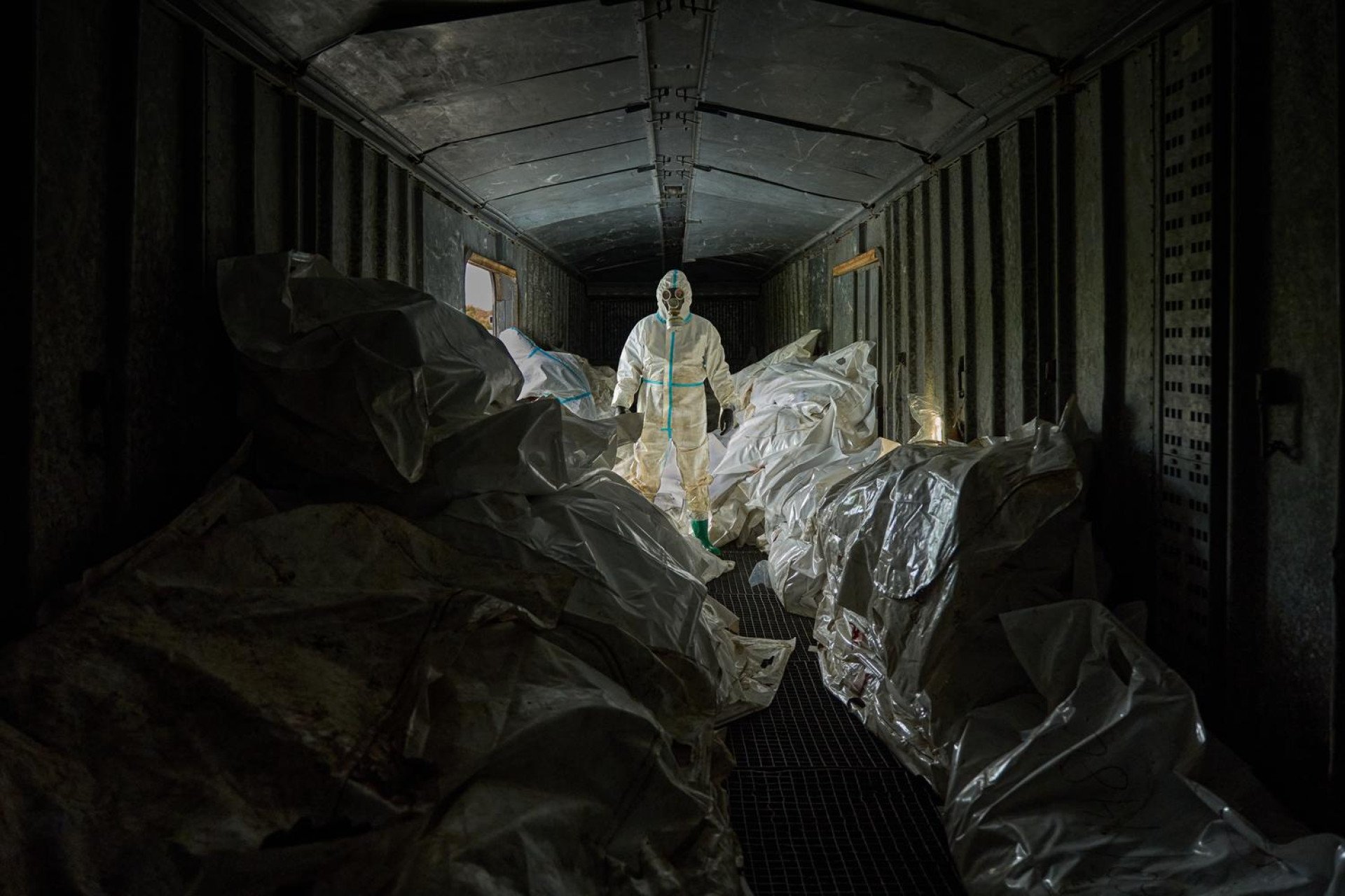 A Ukrainian forensic expert in a white hazmat suit and gas mask stands inside a dark, crowded railway car filled with white body bags of returned dead.