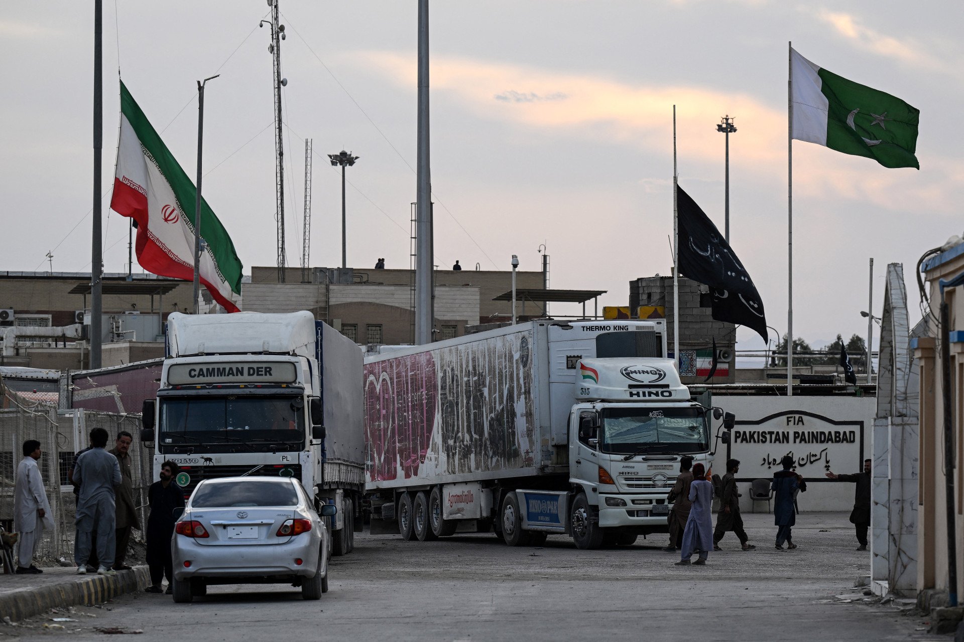 PAKISTAN-IRAN-US-ISRAEL-CONFLICT An Iranian cargo truck crosses into the Pakistan-Iran border as an Iranian national flag (L) flies at half-mast at Taftan, Balochistan province on March 1, 2026 after the death of Iran's supreme leader Ayatollah Ali Khamen