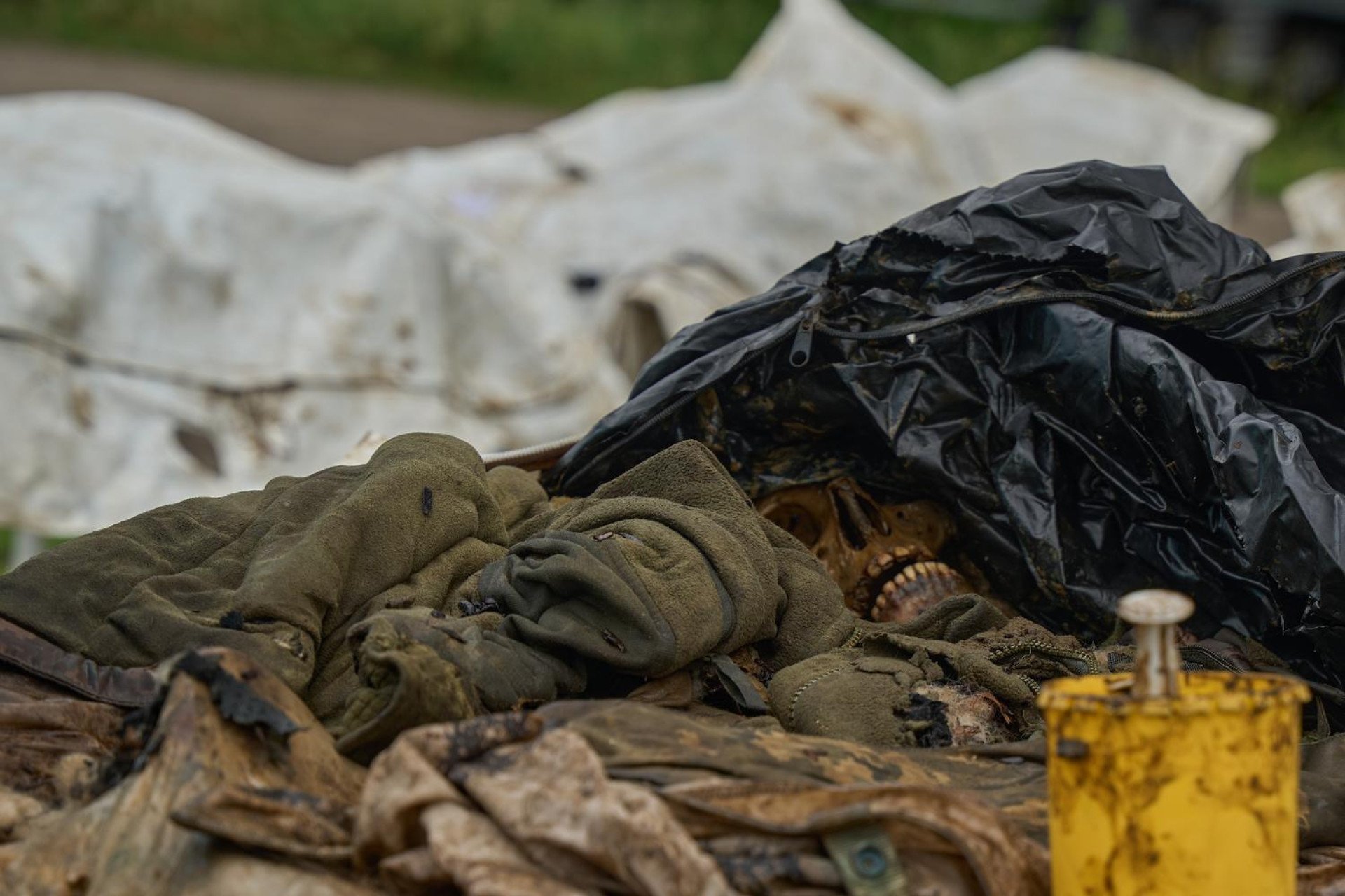 An outdoor, close-up photograph shows a skeletal human skull partially visible beneath a torn black plastic body bag and a bunched-up olive green military fleece jacket. The skull's teeth are prominent, and the surrounding remains are deeply degraded, covered in dirt and organic matter. In the foreground, a stained yellow medical container sits next to piles of decayed uniform fabric, with white body bags blurred in the background.