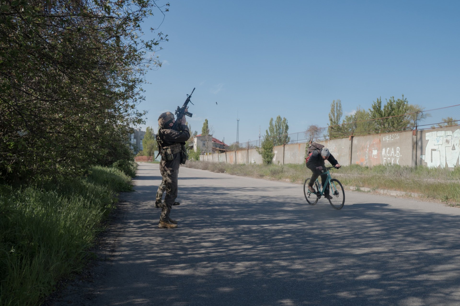 A member of the 34th Marines shoots at a Russian FPV drone as a civilian on a bicycle ducks while passing by. Kherson, Ukraine, May 2026. Photo: Joshua Olley / UNITED24 Media.