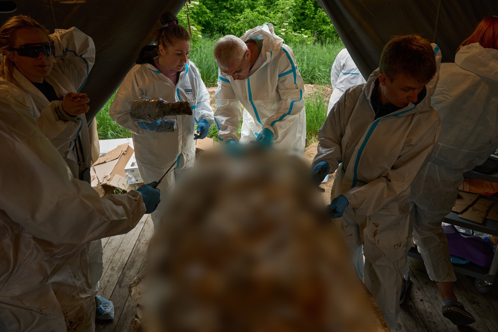 Inside a military field tent, a team of five Ukrainian forensic experts wearing white protective hazmat suits and blue nitrile gloves works around a heavily mummified, decomposed human body laid out on an examination table. In the center, an older male pathologist with glasses carefully inspects the oral and dental structures of the skull, while a female colleague assists from the left. Syringes and small sample containers rest on the chest of the deceased soldier during the post-mortem procedure.