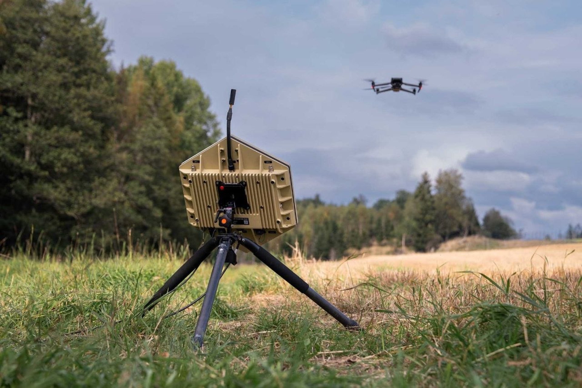 Dispositivo tecnológico militar de color arena con forma poligonal montado en un trípode sobre el césped, apuntando hacia un dron cuadricóptero que vuela bajo un cielo nublado.