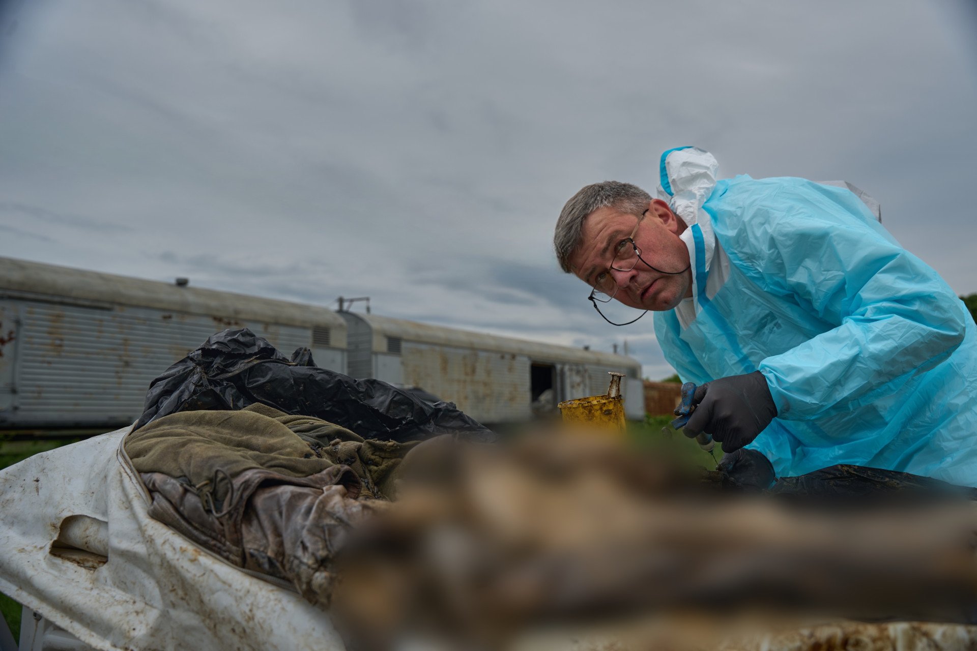 An outdoor photograph shows a middle-aged male Ukrainian forensic expert with glasses, wearing a light blue protective gown over a white hazmat suit and black gloves. He is leaning down to extract a bone marrow or tissue sample with a syringe from highly degraded, mummified remains on an outdoor stretcher. In the background, a gray refrigerated freight train car sits under a cloudy, overcast sky.