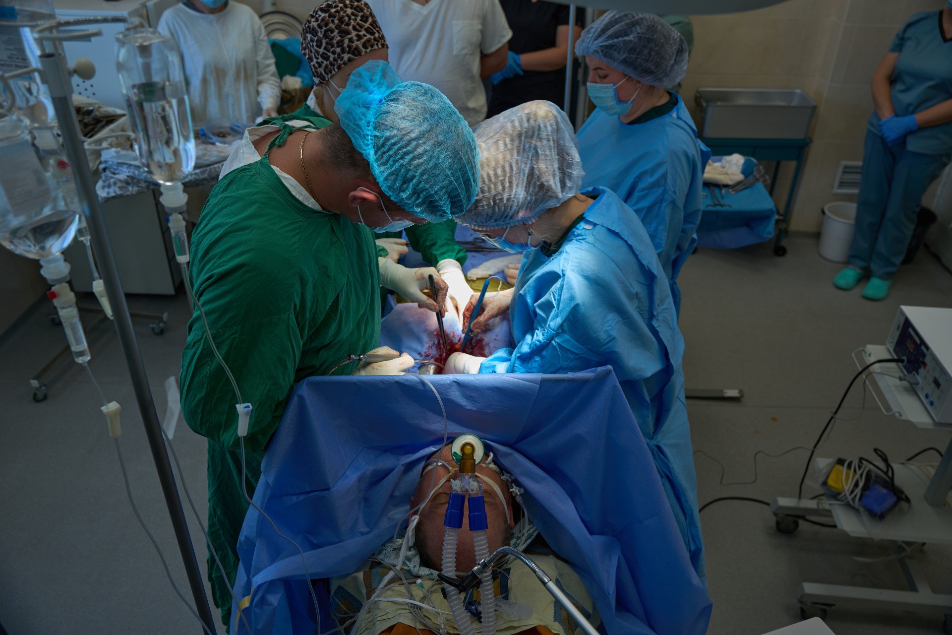 Safely inside the operating theater, the surgical team begins the chest incision to explant the healthy heart. While an anesthesiologist monitors the donor's vital signs behind the sterile drape, the surgeons work with absolute focus. In the high-stakes world of wartime transplantation, this step marks the early stages of a strict seven-hour countdown—the maximum time a donor heart can survive outside the body before being successfully transplanted into the recipient. (Photo: Mykyta Shandyba / UNITED24 Media) Ukrainian surgical team performing open-chest heart explant surgery on a donor patient on a life-support machine in a Kyiv area operating room.