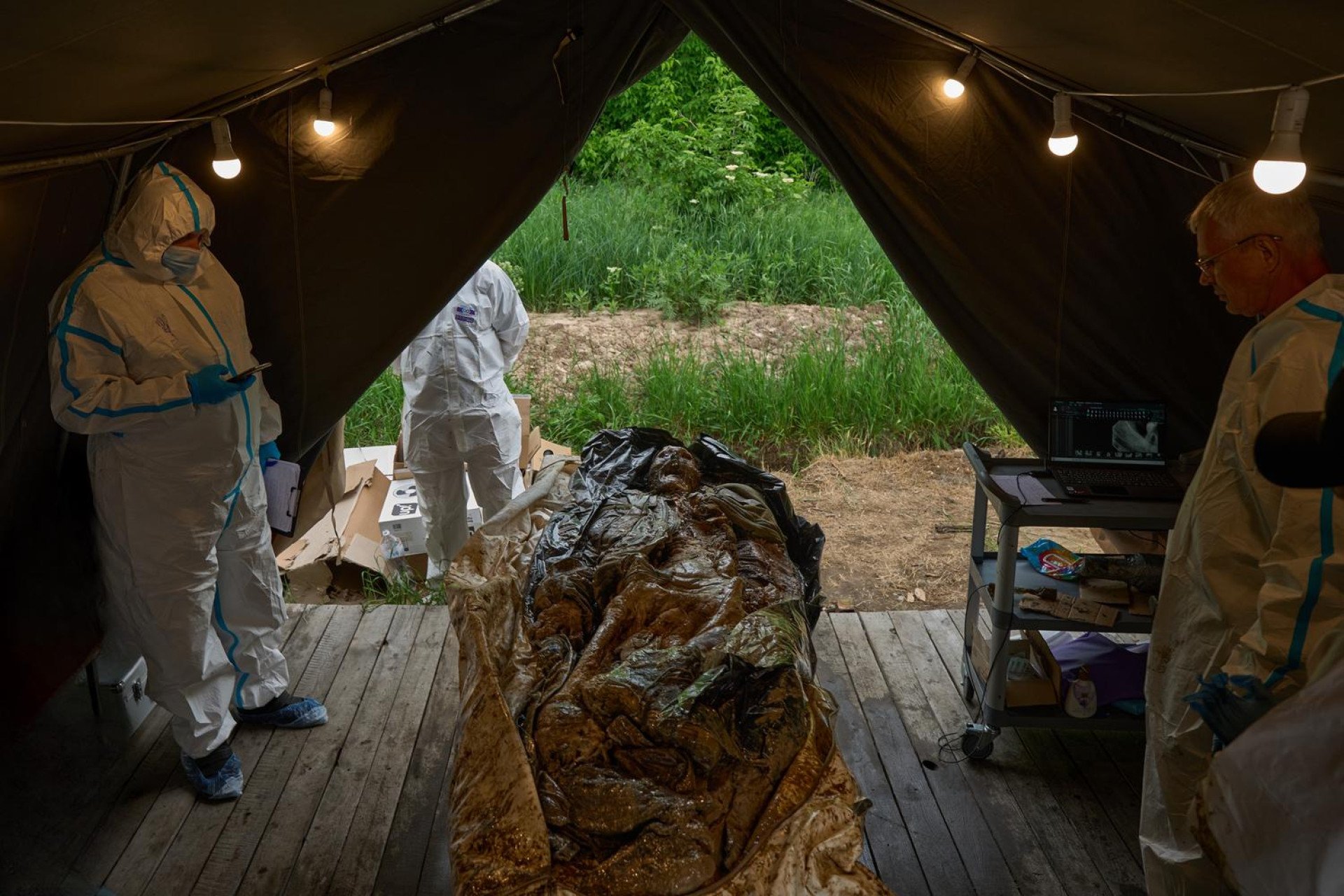 Inside a temporary field tent lit by hanging lightbulbs, Ukrainian forensic experts in white hazmat suits prepare to work on heavily decomposed human remains laid on a wooden floor. One expert stands on the left holding a smartphone and a clipboard, while another stands on the right near a mobile cart equipped with a laptop displaying a forensic image or scan. The open back of the tent looks out onto a green, grassy outdoor area.