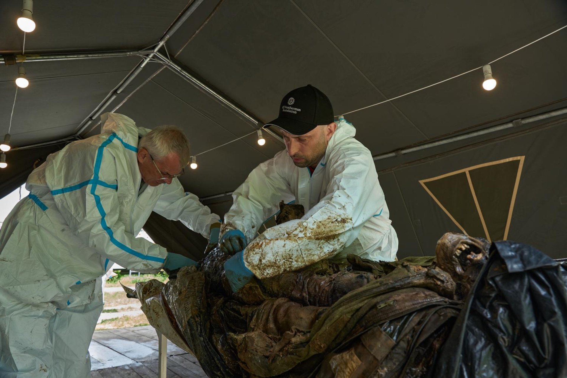 Inside a temporary tent station lit by incandescent bulbs, two male Ukrainian forensic experts in white hazmat suits and blue protective gloves work together on highly decomposed, mummified human remains. The expert on the right wears a black baseball cap with "RT Weatherman Foundation" logo and leans closely over the upper torso of the body. The older expert on the left with glasses assists with the delicate post-mortem examination.