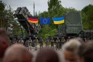 Ukrainian service members stand in front of a Patriot air defense system alongside German, EU, and Ukrainian flags. Ukrainian service members stand in front of a Patriot air defense system alongside German, EU, and Ukrainian flags.