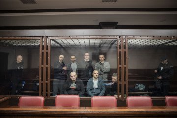 The image shows a group of individuals in a courtroom, presumably on trial, as they stand within a glass-enclosed dock. (Photo: Mediazona)