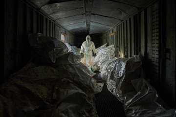 A Ukrainian forensic expert in a white hazmat suit and gas mask stands inside a dark, crowded railway car filled with white body bags of returned dead.