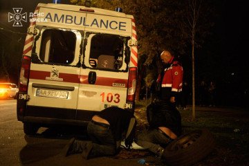 An ambulance with its rear windows blown out after a Russian missile strike in Kyiv at night, as a paramedic stands beside it and two people kneel at the back of the vehicle.