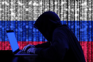 Un hacker con una sudadera oscura con capucha trabaja en una computadora portátil frente a un fondo digital con la bandera rusa y código binario. Foto ilustrativa. (Fuente: Getty Images)