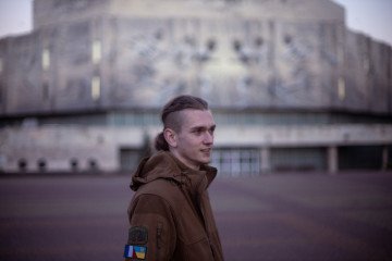 Un joven con una chaqueta marrón de estilo militar con parches ucranianos está de pie al aire libre, sonriendo levemente, con un gran edificio de hormigón borroso al fondo.