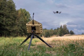 Dispositivo tecnológico militar de color arena con forma poligonal montado en un trípode sobre el césped, apuntando hacia un dron cuadricóptero que vuela bajo un cielo nublado.