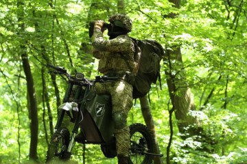 A Ukrainian special forces soldier uses binoculars while standing on a WolfStorm electric bike in a dense forest. The bike’s low noise and thermal signature support covert reconnaissance missions. (Source: UkrSpecConsulting)