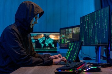 Anonymous computer hacker in white mask and hoodie. Obscured dark face using laptop computer for cyber attack and calling on cellphone. Illustrative photo. (Source: Getty Images)