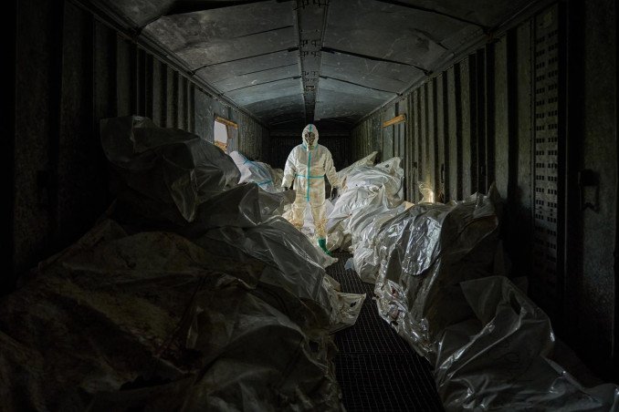 A Ukrainian forensic expert in a white hazmat suit and gas mask stands inside a dark, crowded railway car filled with white body bags of returned dead.