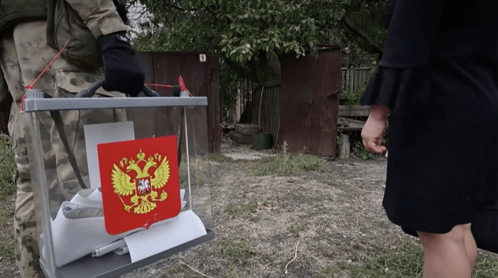 A resident casts a ballot during Russian-organized early voting in the Russian-occupied Donetsk region as an armed serviceman stands beside a mobile ballot box, September 14, 2026. 