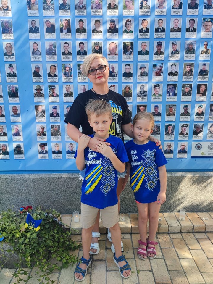 Maryna with her children, Myroslav and Solomiia, beside Vasyl’s photo on Kyiv’s Wall of Remembrance for fallen Ukrainian soldiers. (Source: Maryna's personal archive.)