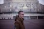 A young man in a brown military-style jacket with Ukrainian patches stands outdoors, slightly smiling, with a large, blurred concrete building in the background.